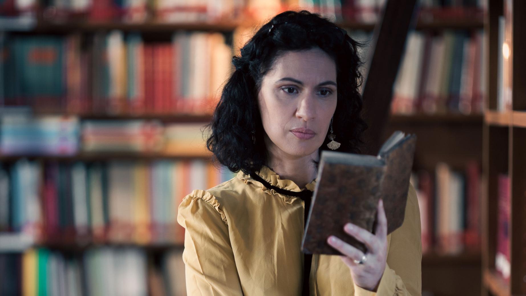 A woman stands in a historic library filled with bookshelves and long wooden tables covered in documents, viewed through a ZEISS lens.