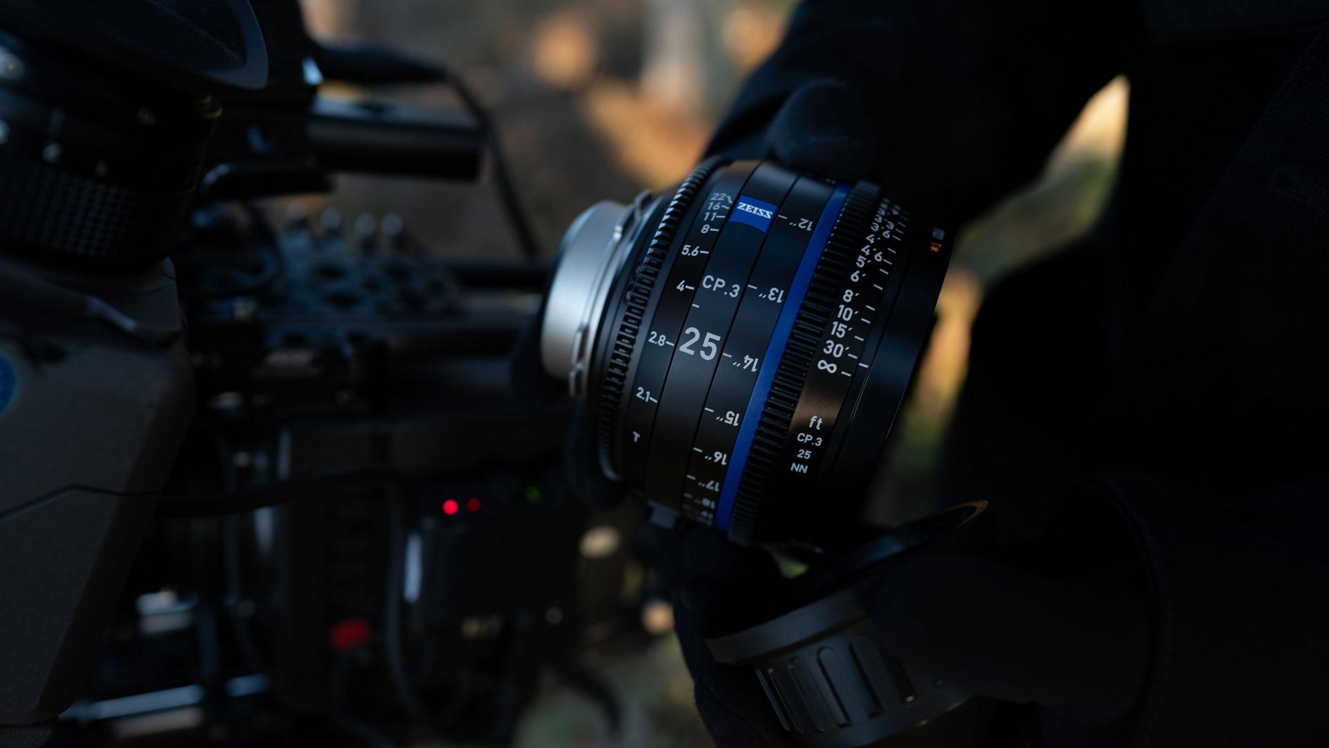 A person is holding a ZEISS 25mm cinema lens next to a professional video camera setup.