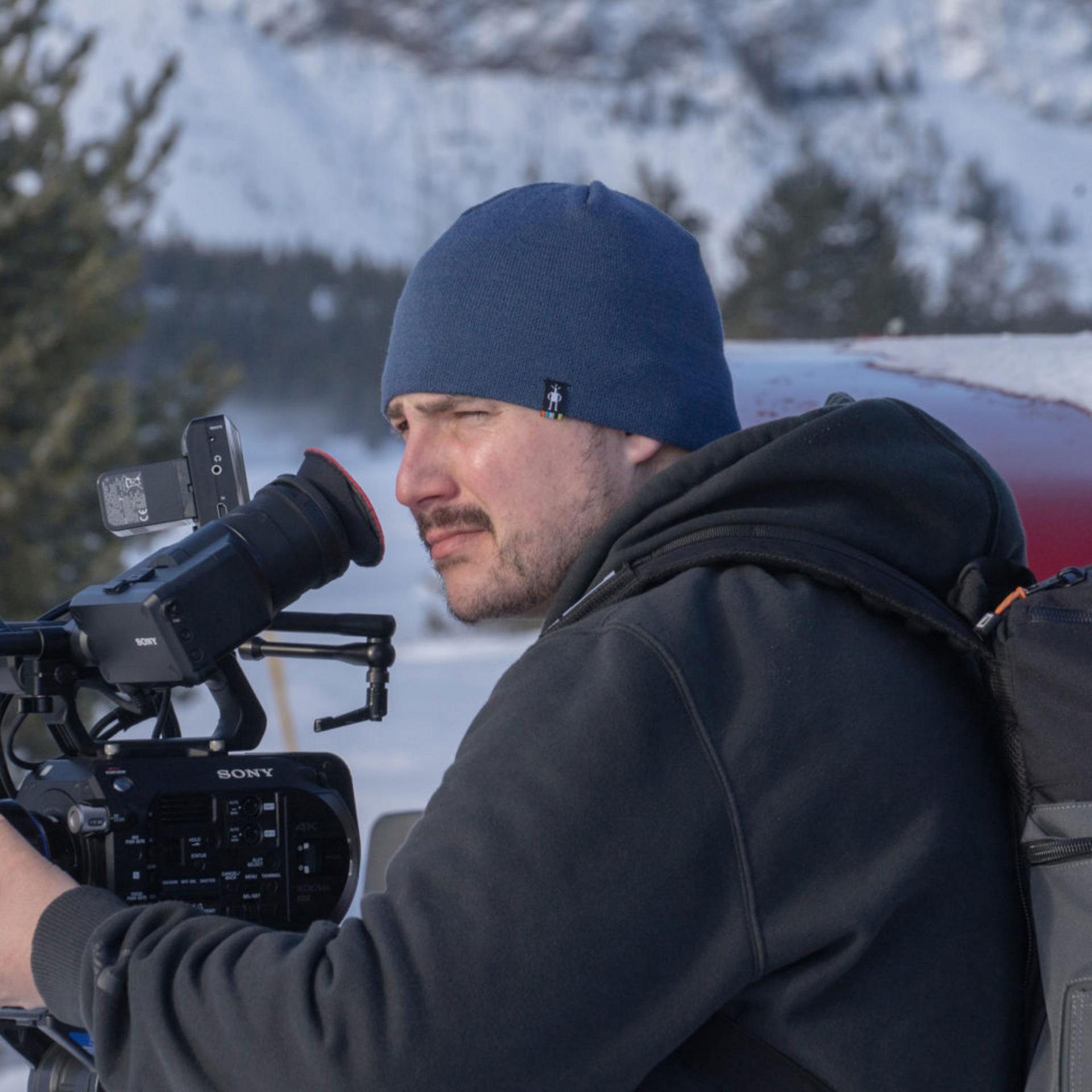Cameraman filming outdoors in a snowy mountain setting using a SONY camera equipped with a ZEISS lens.