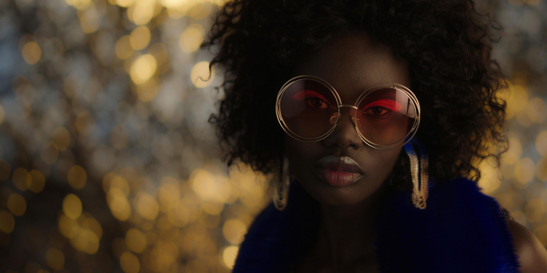 Woman wearing oversized round ZEISS eyewear with tinted lenses, standing in front of a softly lit, bokeh-filled background.