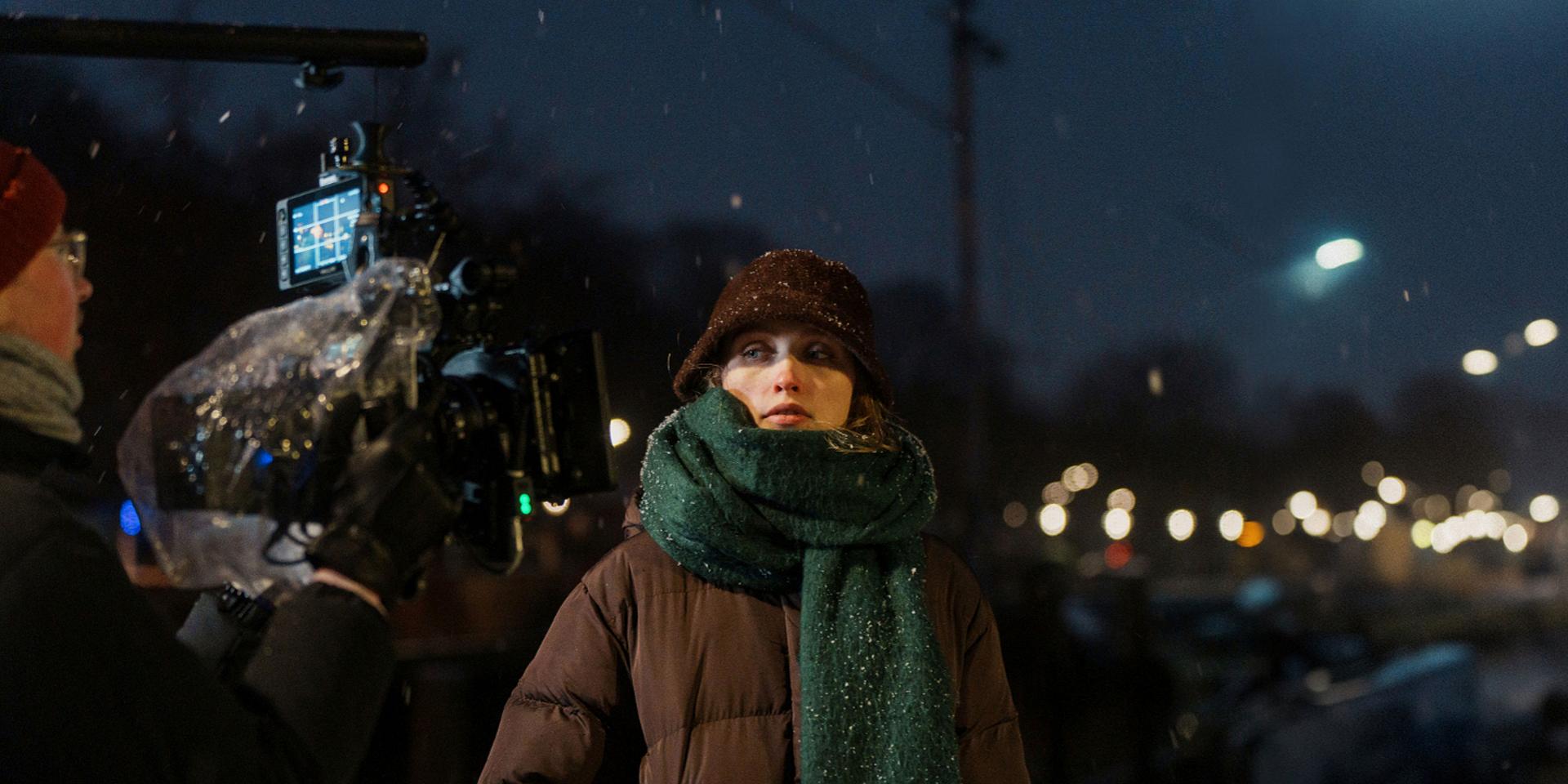 A film crew captures a scene at night featuring a woman in winter clothing, illuminated by streetlights and light snowfall, using a professional camera setup with ZEISS lenses.