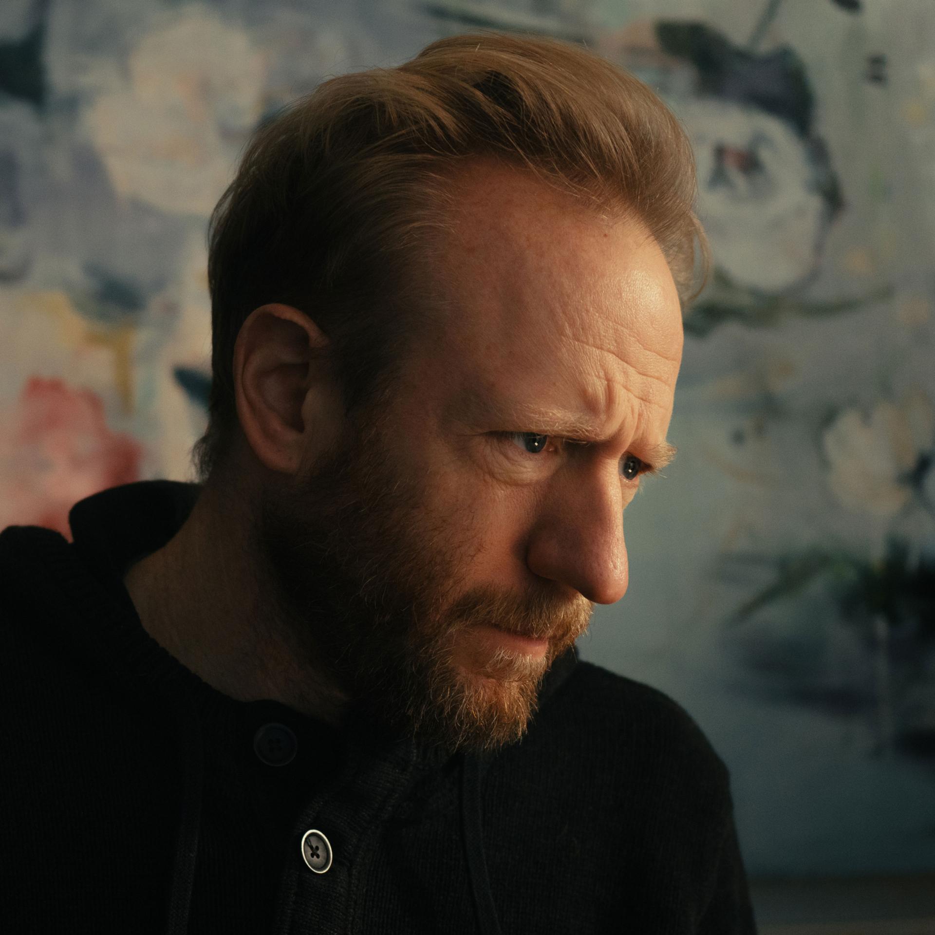 A bearded man in a dark sweater is looking thoughtfully to the side in front of a softly blurred, artistic background, captured with ZEISS-quality clarity.