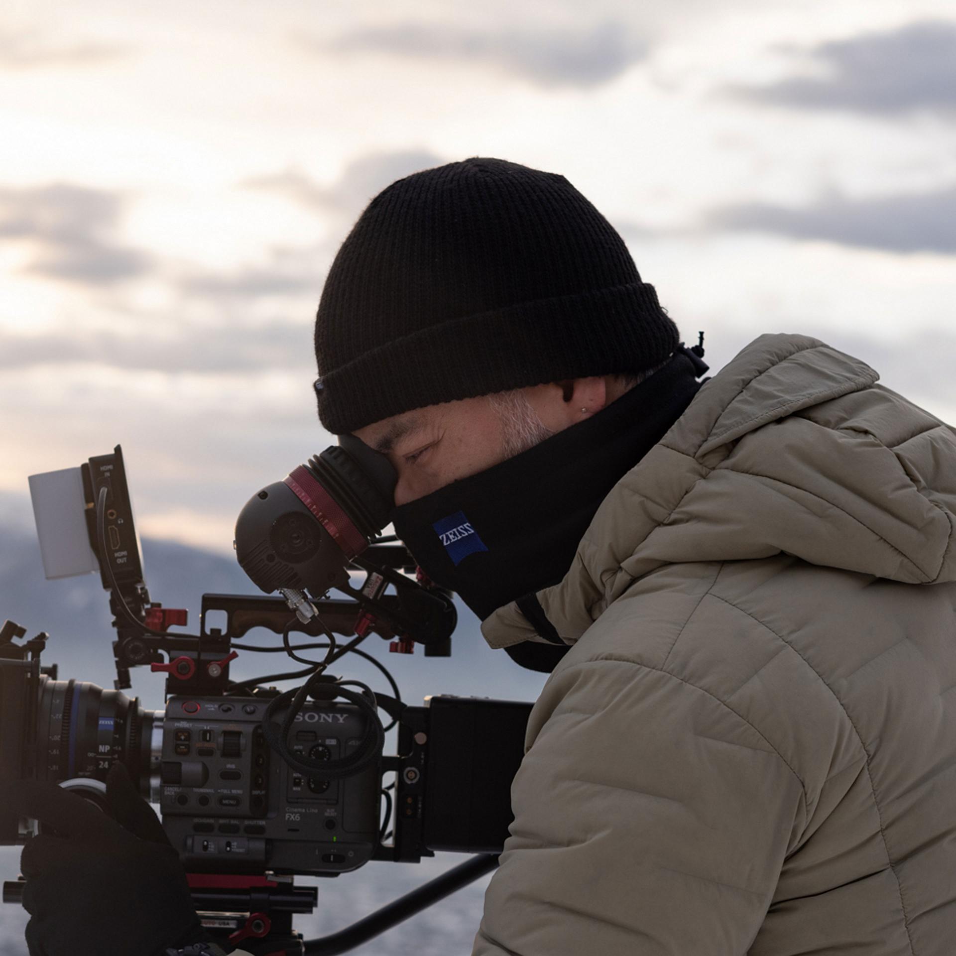 A person wearing winter clothing is operating a professional video camera with a ZEISS branded eyepiece in an outdoor setting.