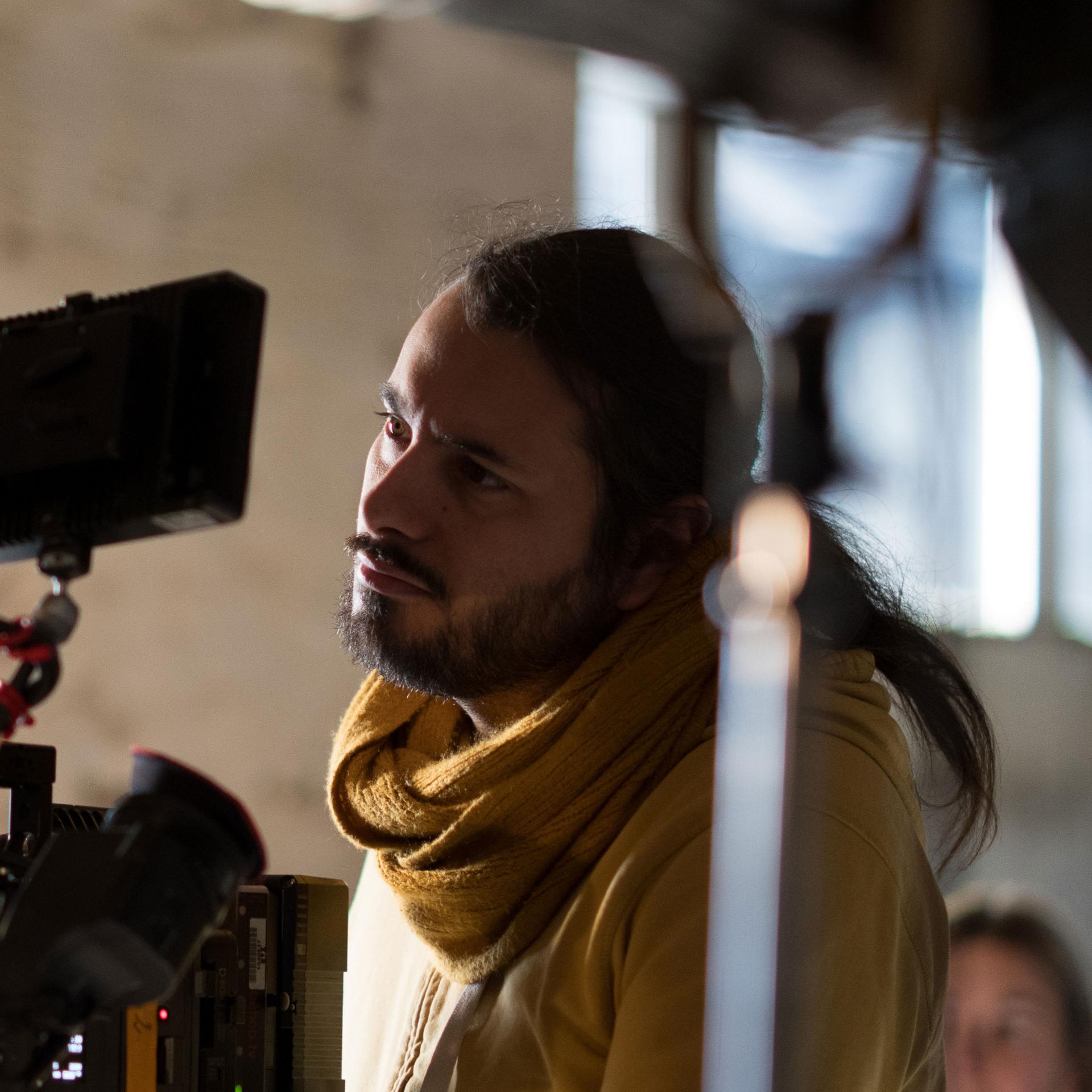 A man wearing a yellow scarf closely observes a monitor on a professional film set with ZEISS camera equipment.