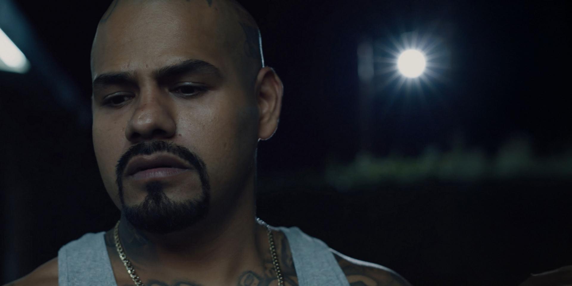 A man with a shaved head and goatee is looking down thoughtfully at night, illuminated by a bright light source in the background.