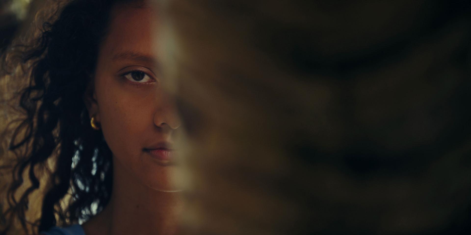 A woman with curly hair is partially visible, with half of her face obscured by a blurred foreground object.