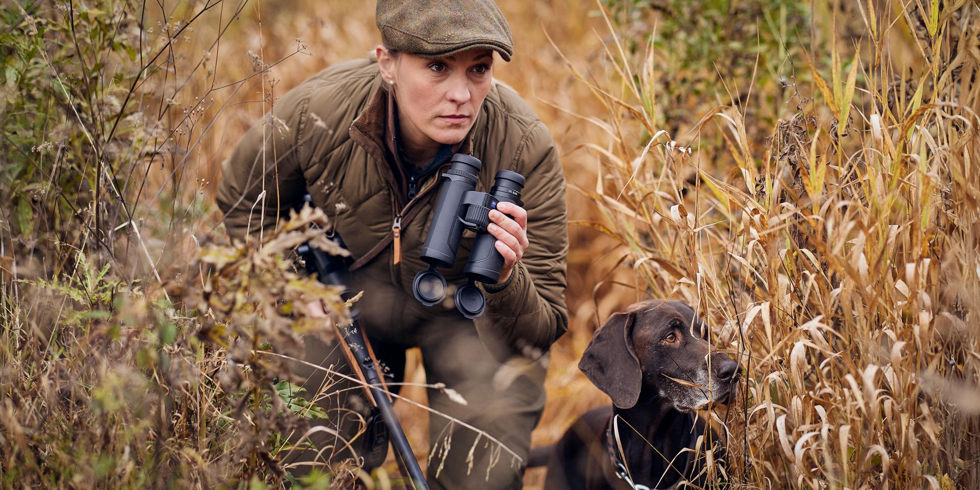 Person in outdoor gear holding ZEISS binoculars beside a hunting dog in tall, dry grass.