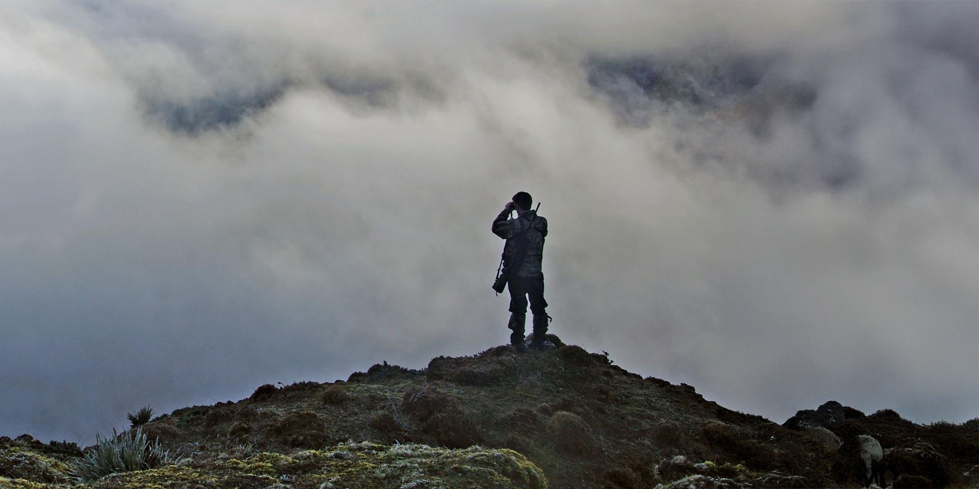 A person stands on a rocky hilltop looking through binoculars under a cloudy sky, highlighting ZEISS optical equipment in use.