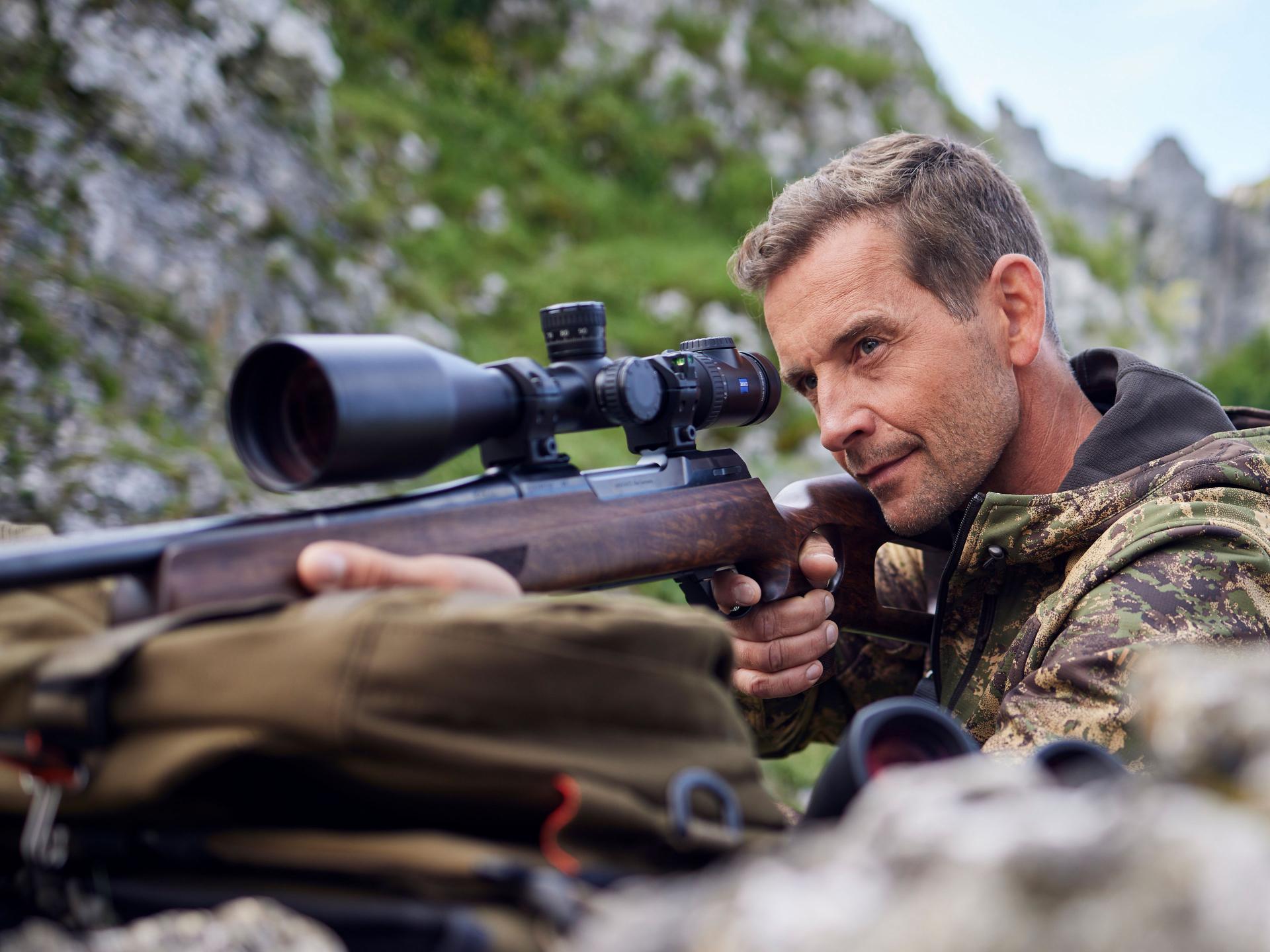 A hunter lies in the mountains aiming through a ZEISS V8 riflescope, showcasing accuracy, versatility, and performance in demanding alpine terrain.