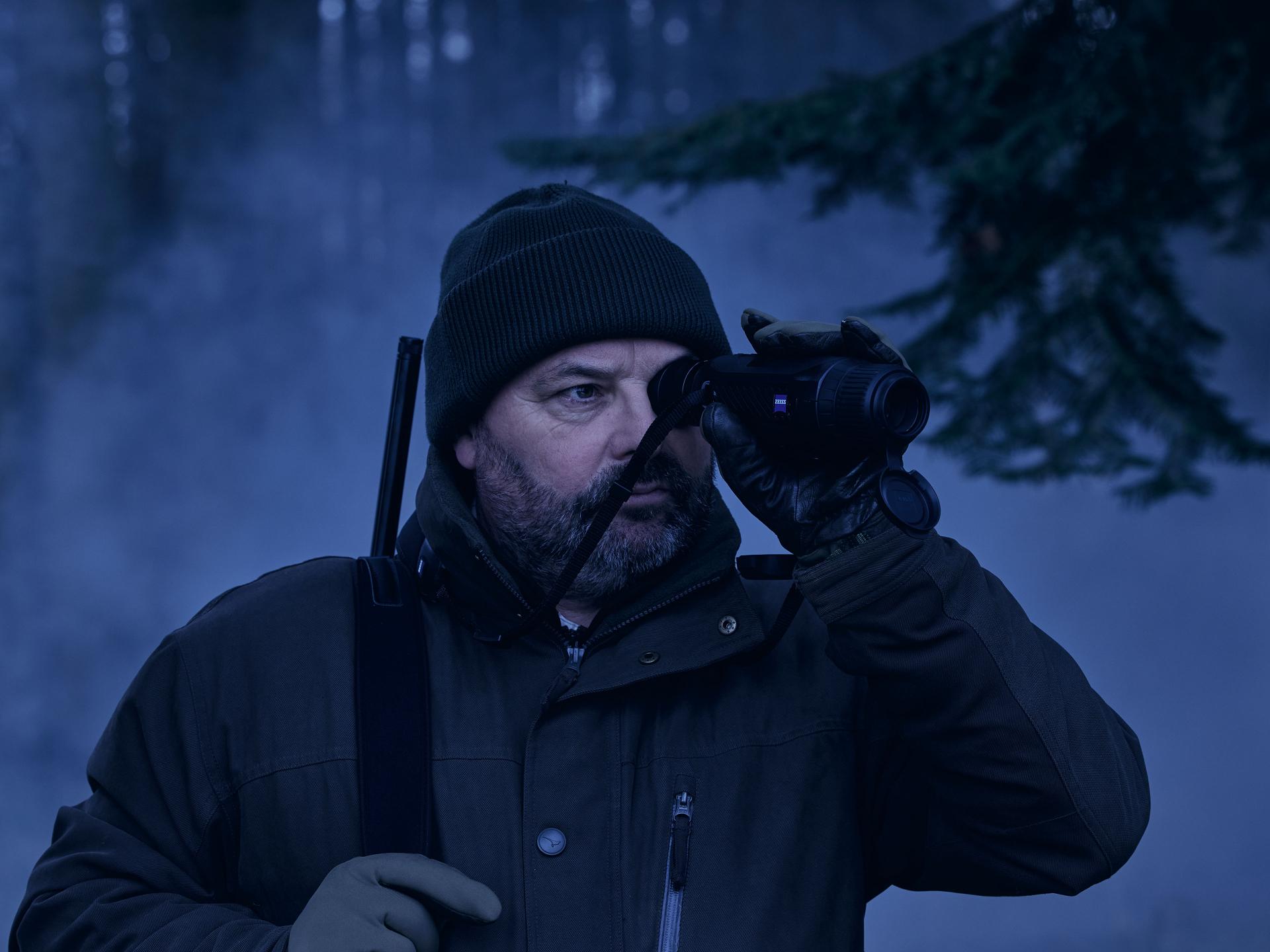 Man in outdoor winter gear using a ZEISS thermal imaging device in a foggy forest setting.