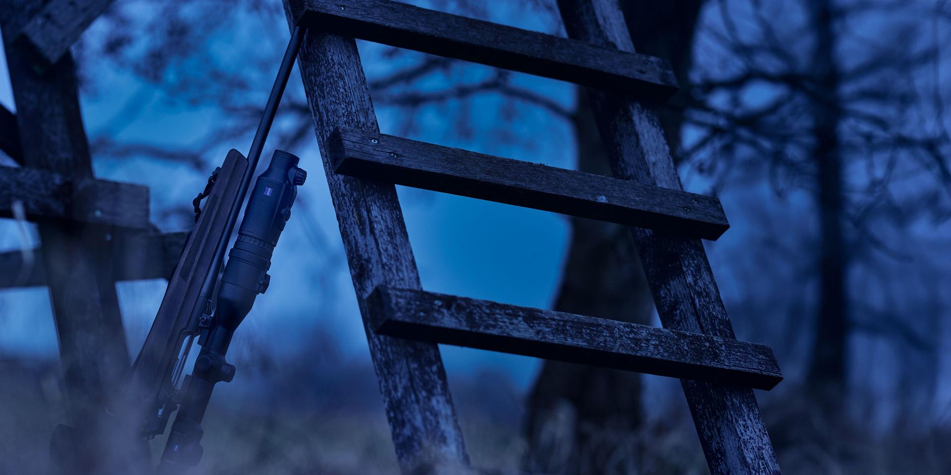 A hunting rifle equipped with a scope is leaning against a wooden ladder in a forest setting at dusk, with ZEISS optics visible.