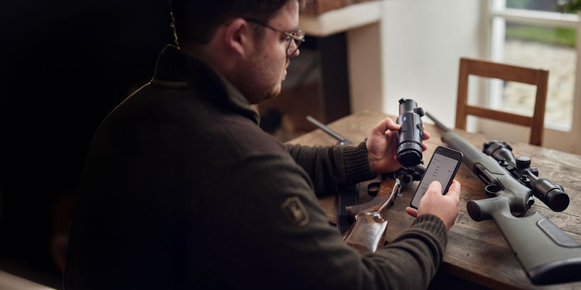 A man sits at a wooden table inspecting a ZEISS riflescope while holding a smartphone, with rifles and another ZEISS optic beside him.