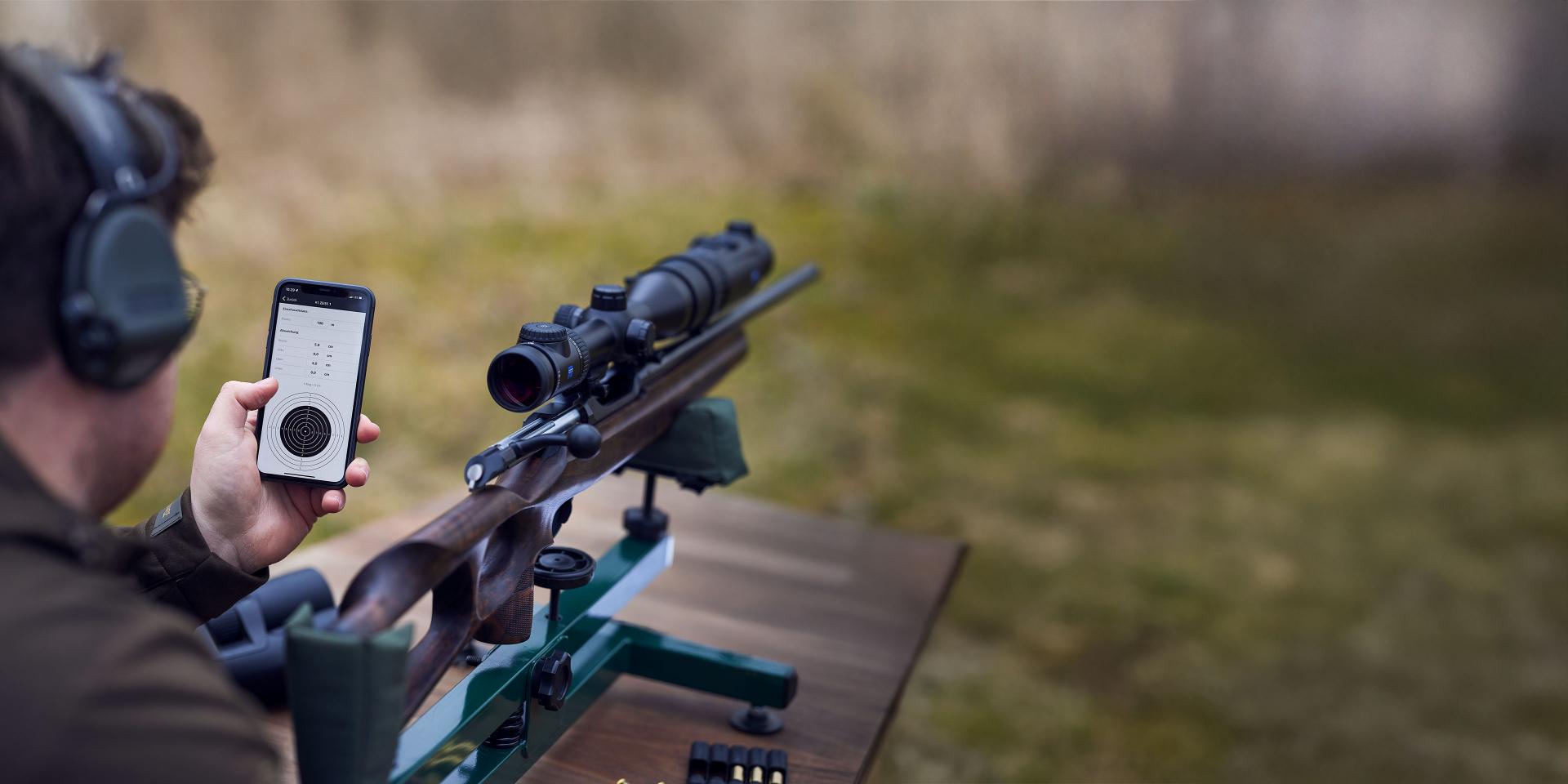 A person at an outdoor shooting range holds a smartphone displaying a ballistic app next to a rifle equipped with a ZEISS riflescope.