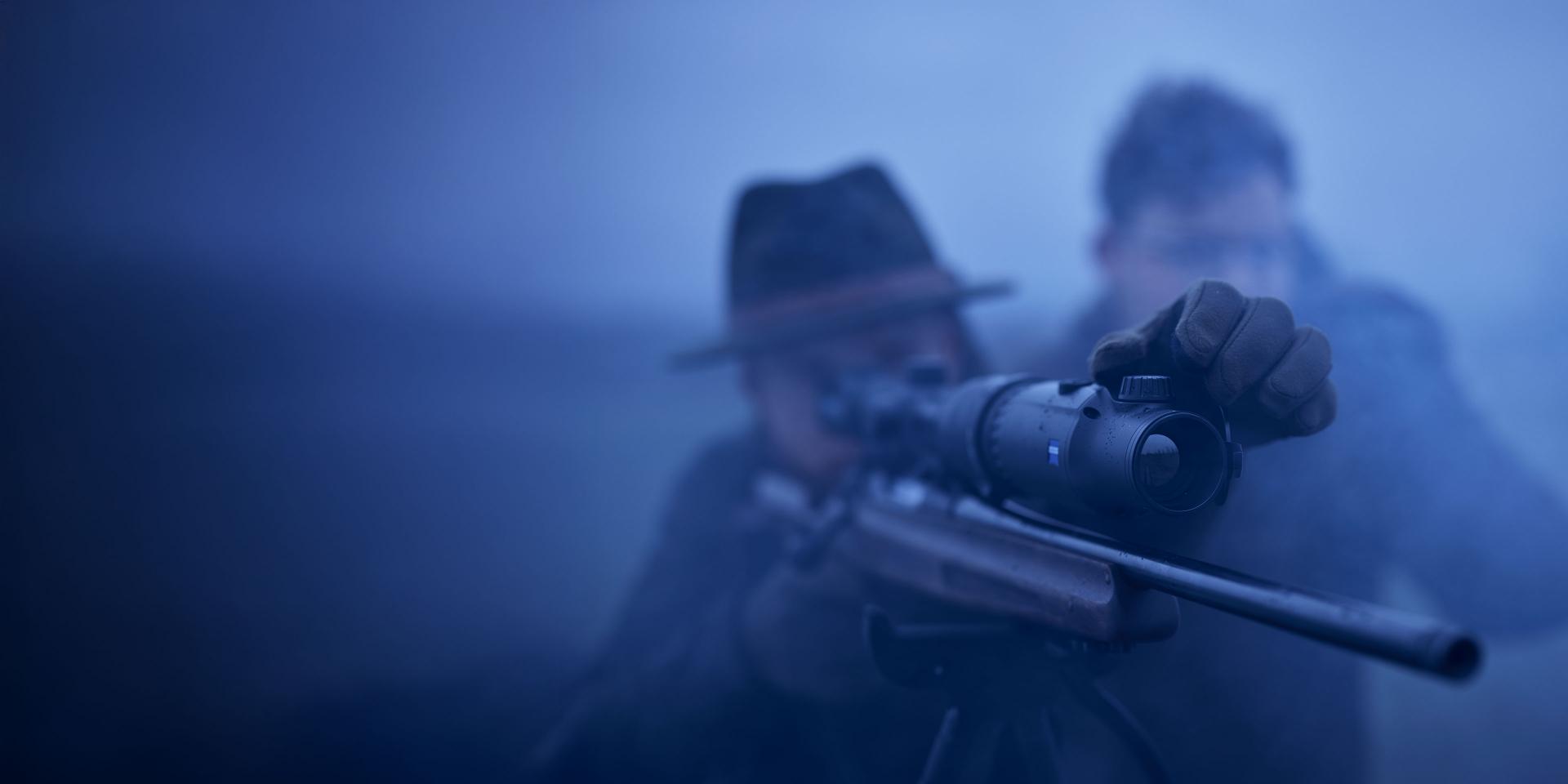 Two people in outdoor gear are aiming a rifle equipped with a ZEISS riflescope in low-light conditions.