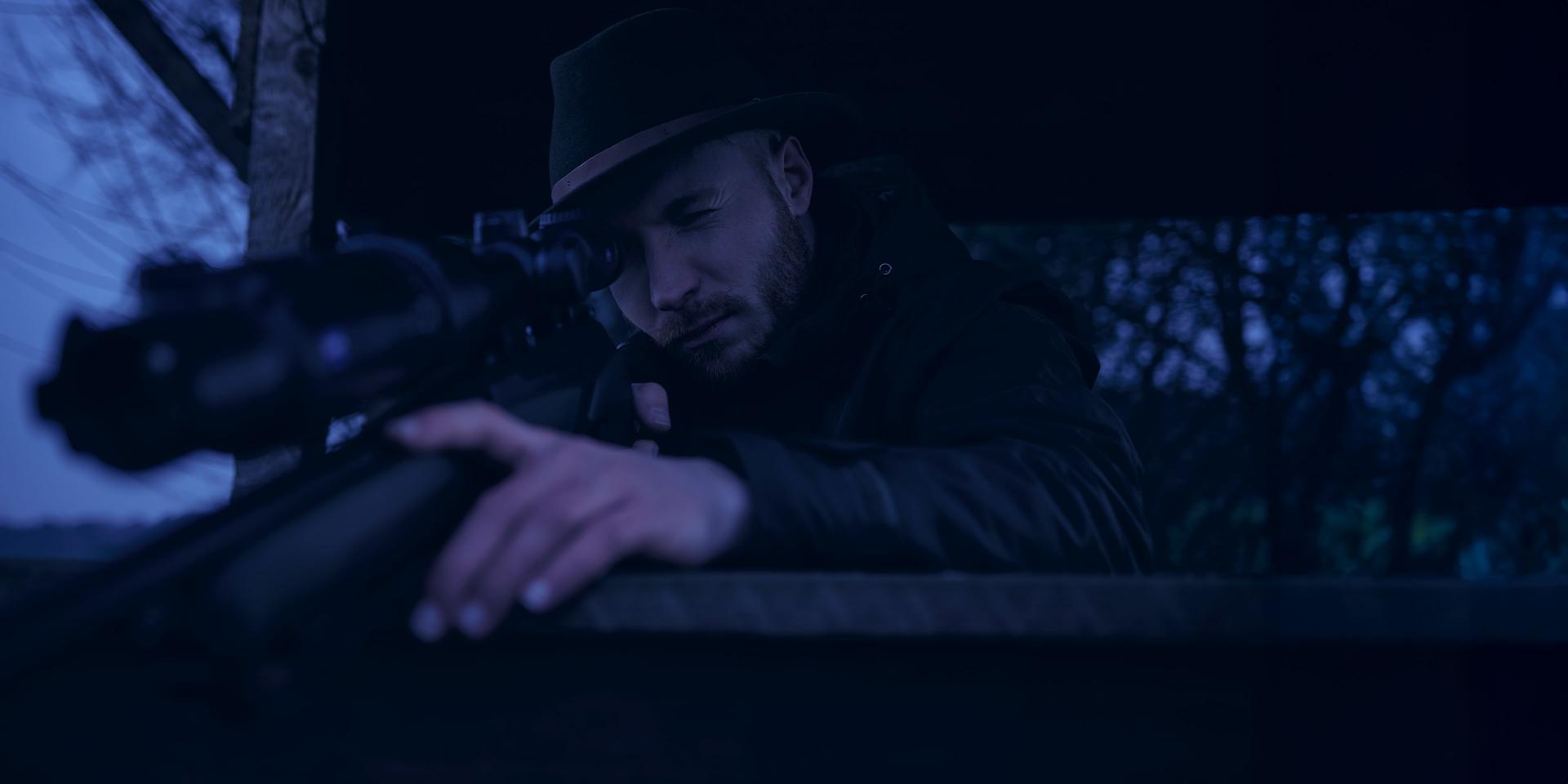 A man wearing a hat is aiming through a ZEISS riflescope in a dark outdoor setting.