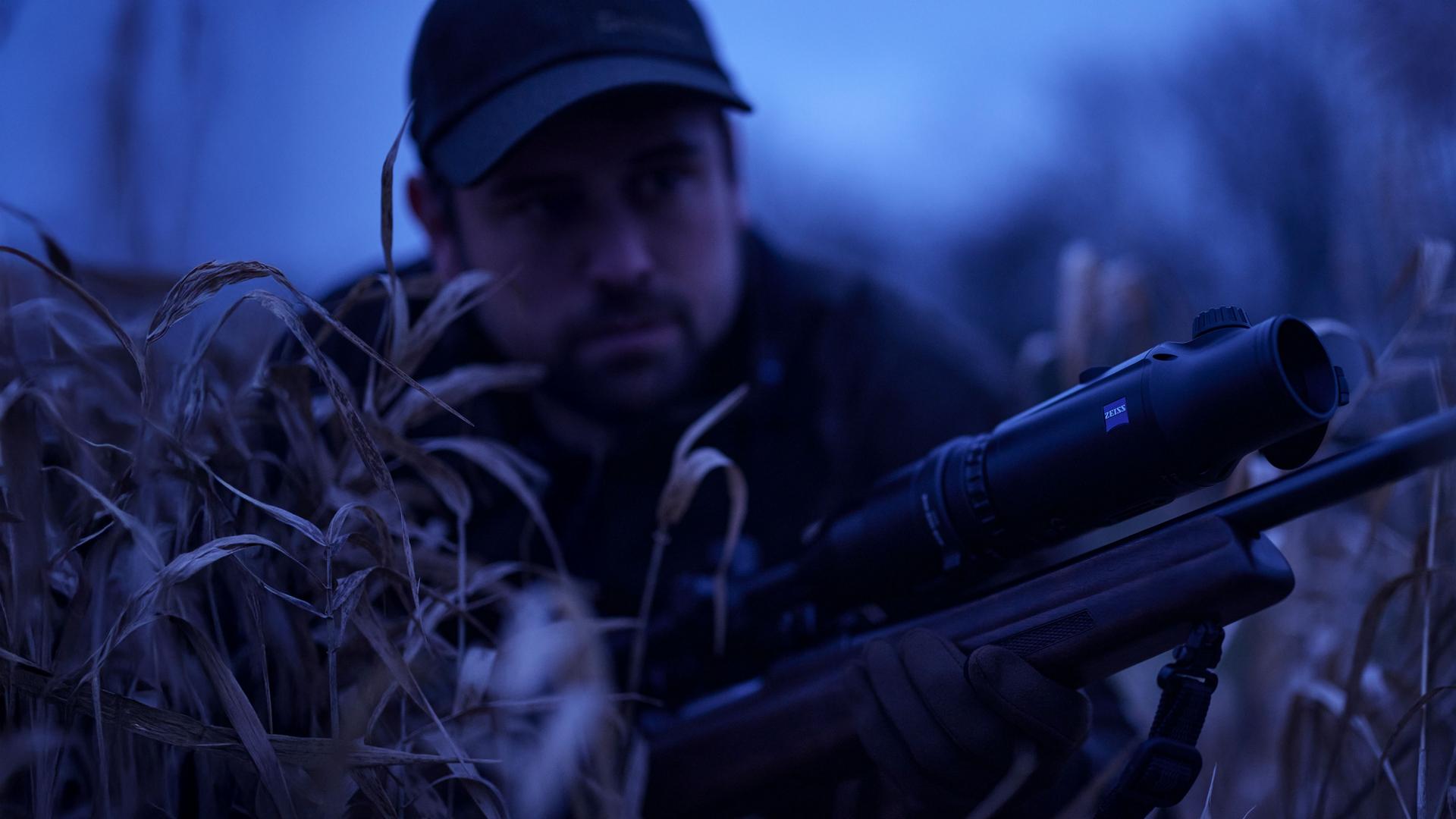 A man in camouflage gear is lying in tall grass at dusk, aiming a rifle equipped with a ZEISS optic.
