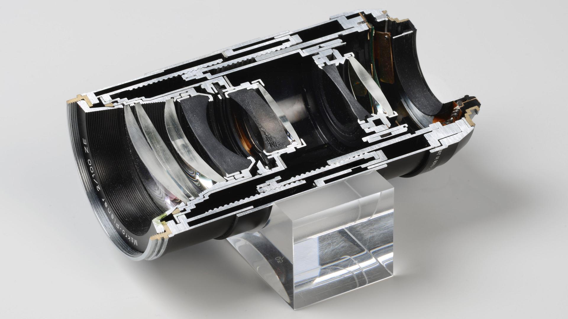 Cross-sectional view of a ZEISS camera lens showing internal optical and mechanical components, mounted on a clear display stand.