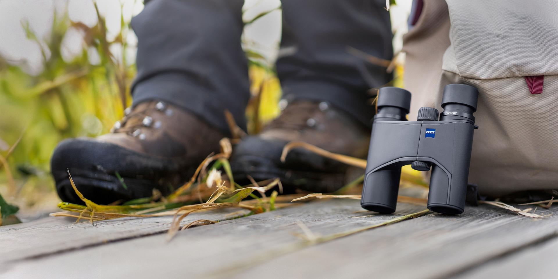 A pair of ZEISS binoculars rests on a wooden surface next to a beige backpack and hiker's boots in an outdoor setting.