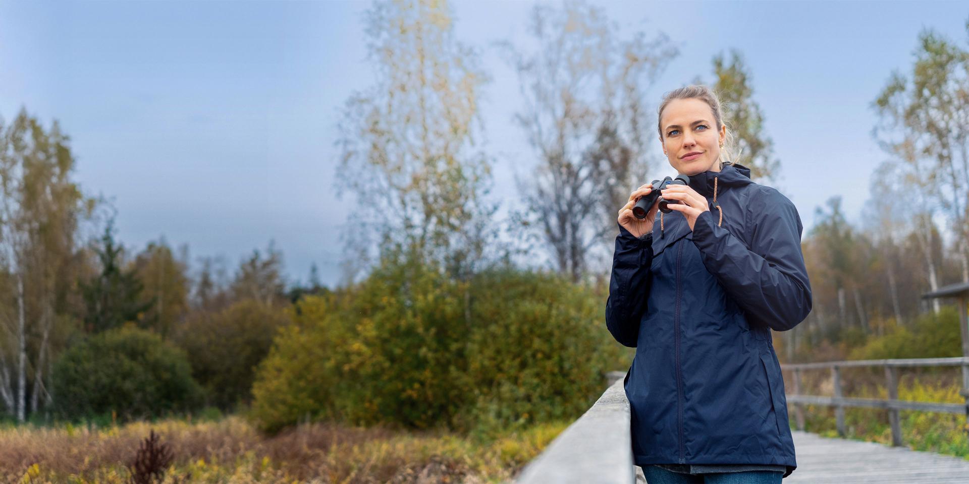 A woman in a navy jacket stands on a wooden path in a natural landscape holding ZEISS binoculars.