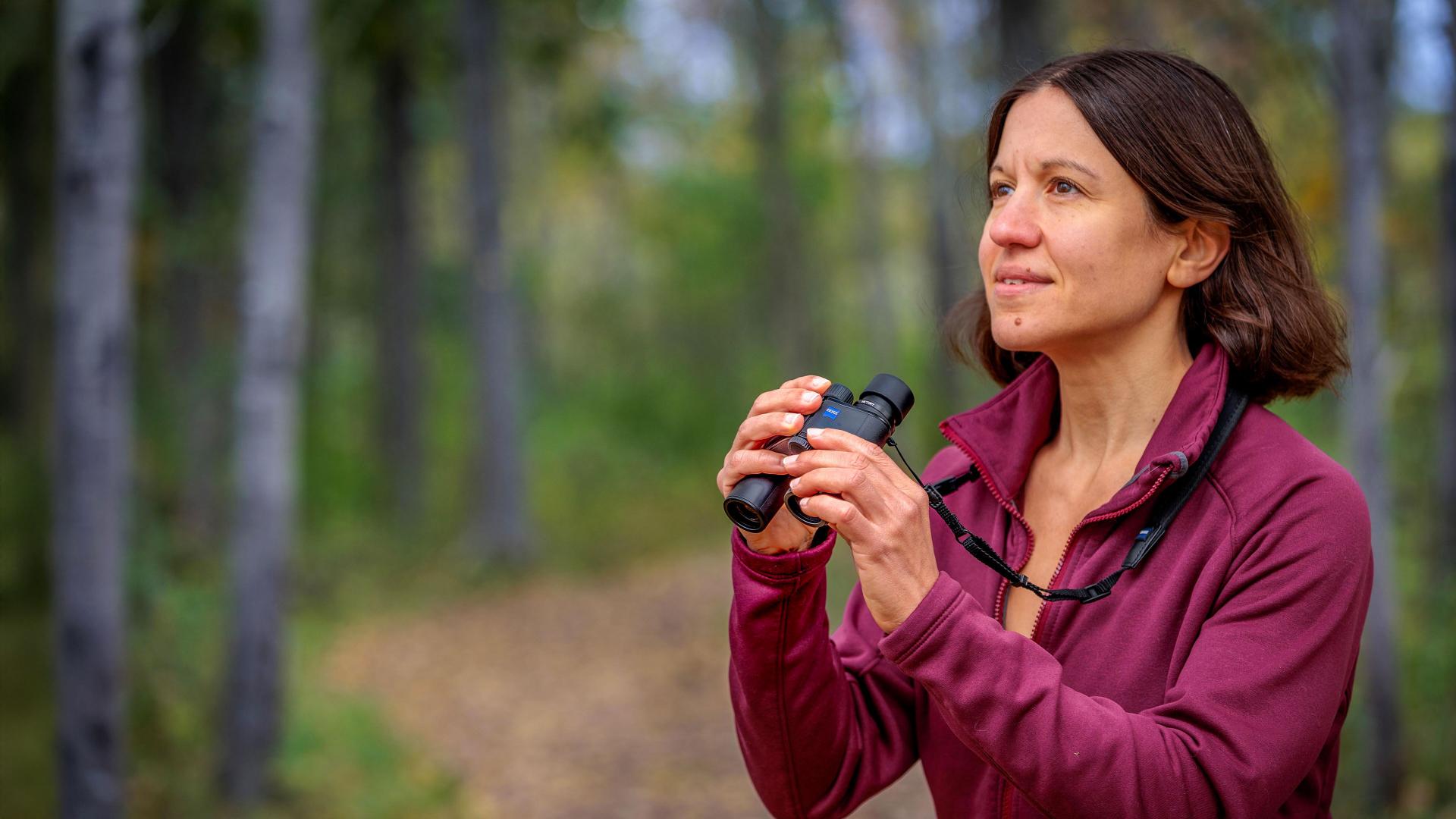 A woman stands on a forest path holding a pair of ZEISS binoculars, looking into the distance.