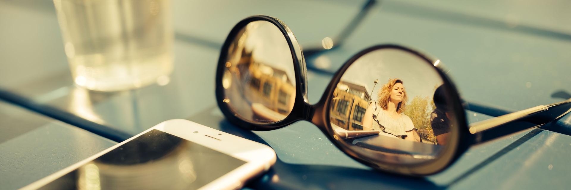 Sunglasses with ZEISS lenses lying on a blue table, reflecting a smiling woman, next to a smartphone and a glass of water.