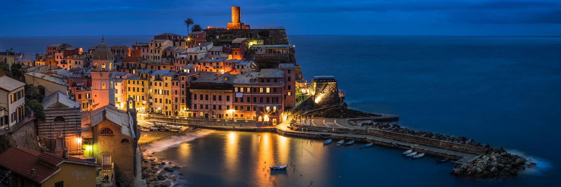 Coastal town with colorful buildings illuminated at night, reflecting in the calm harbor under a deep blue sky, captured in sharp detail.