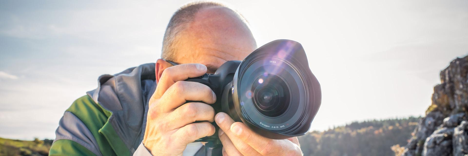 A man is taking a photo outdoors with a DSLR camera equipped with a ZEISS lens.