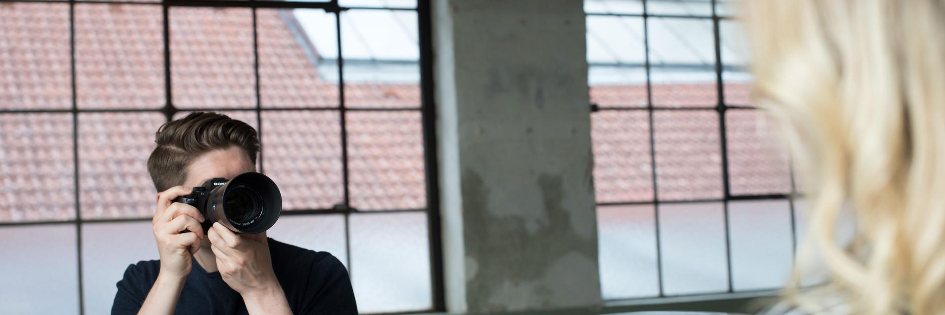 A man is taking a photo of a woman using a ZEISS camera lens in an industrial-style indoor setting.