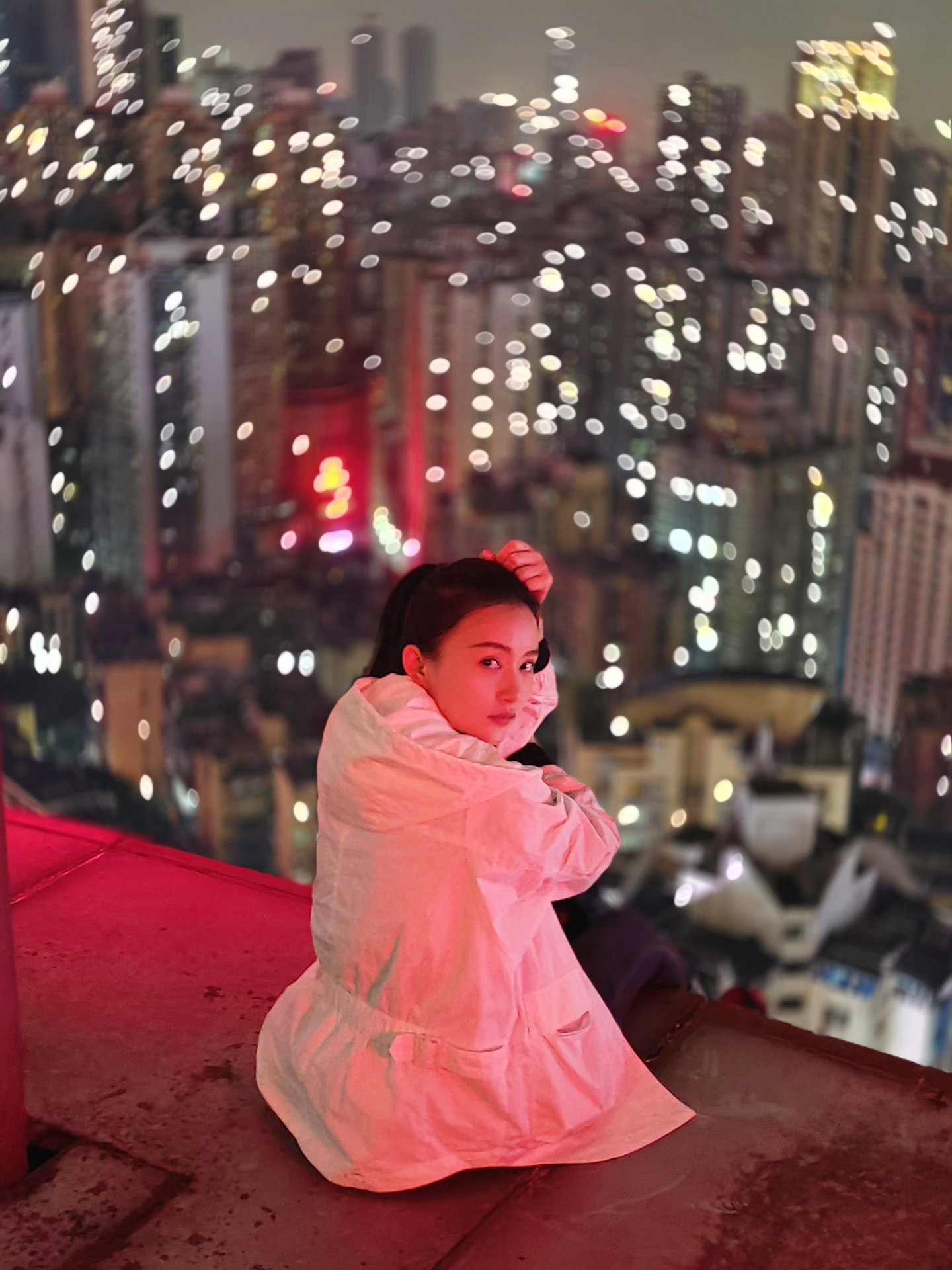 A woman in a white coat sits on a rooftop ledge at night with a vibrant, bokeh-filled cityscape in the background, showcasing ZEISS lens clarity and depth.