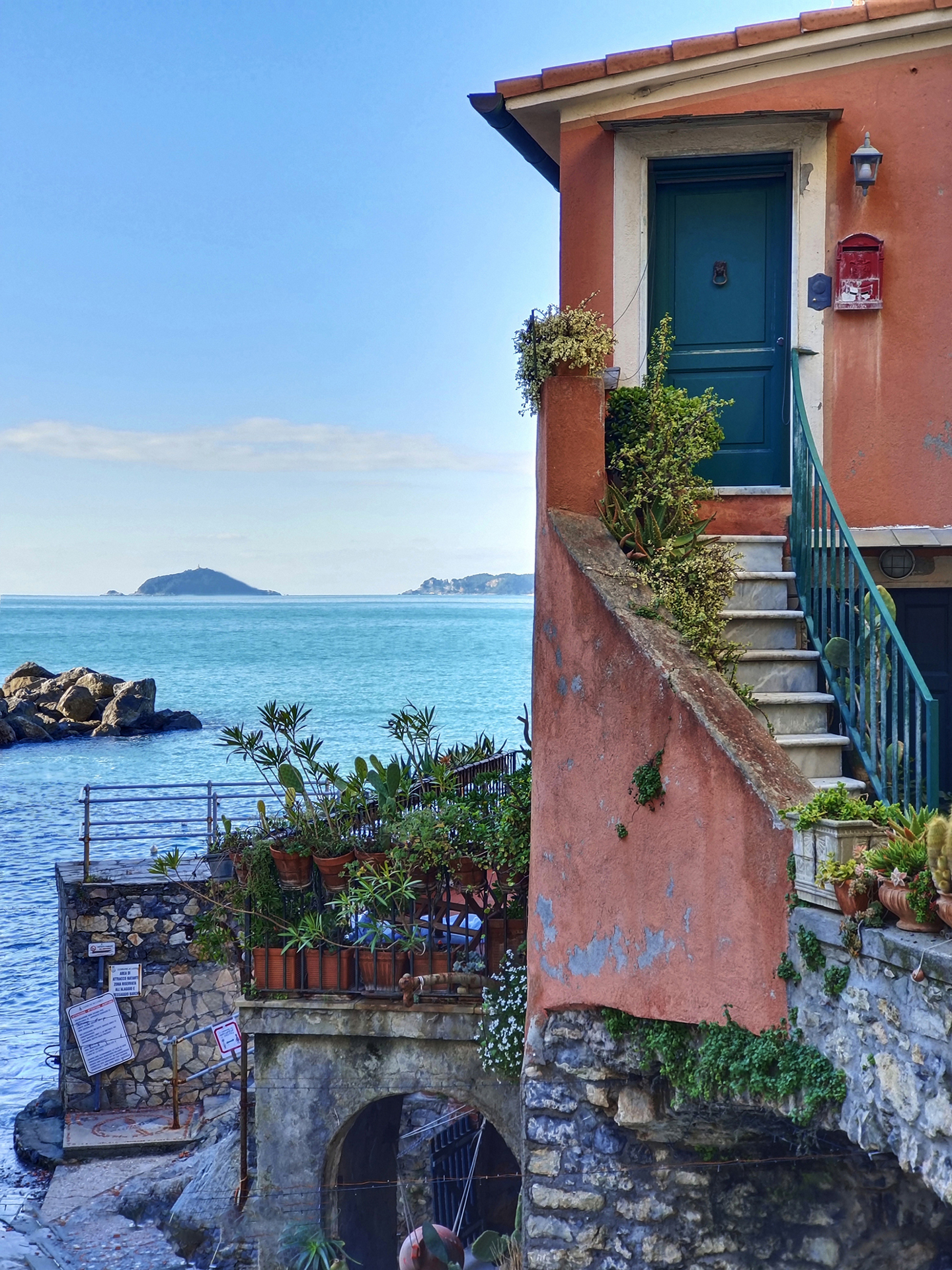 A seaside house with a terracotta exterior, stone steps, and potted plants overlooks a calm blue ocean under a clear sky, captured in vivid detail by ZEISS optics.