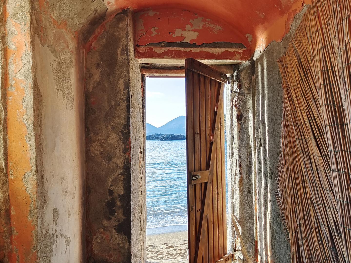 A rustic doorway with a partially open wooden door reveals a serene beach and calm sea with mountains in the distance, viewed from inside a weathered, sand-covered room.