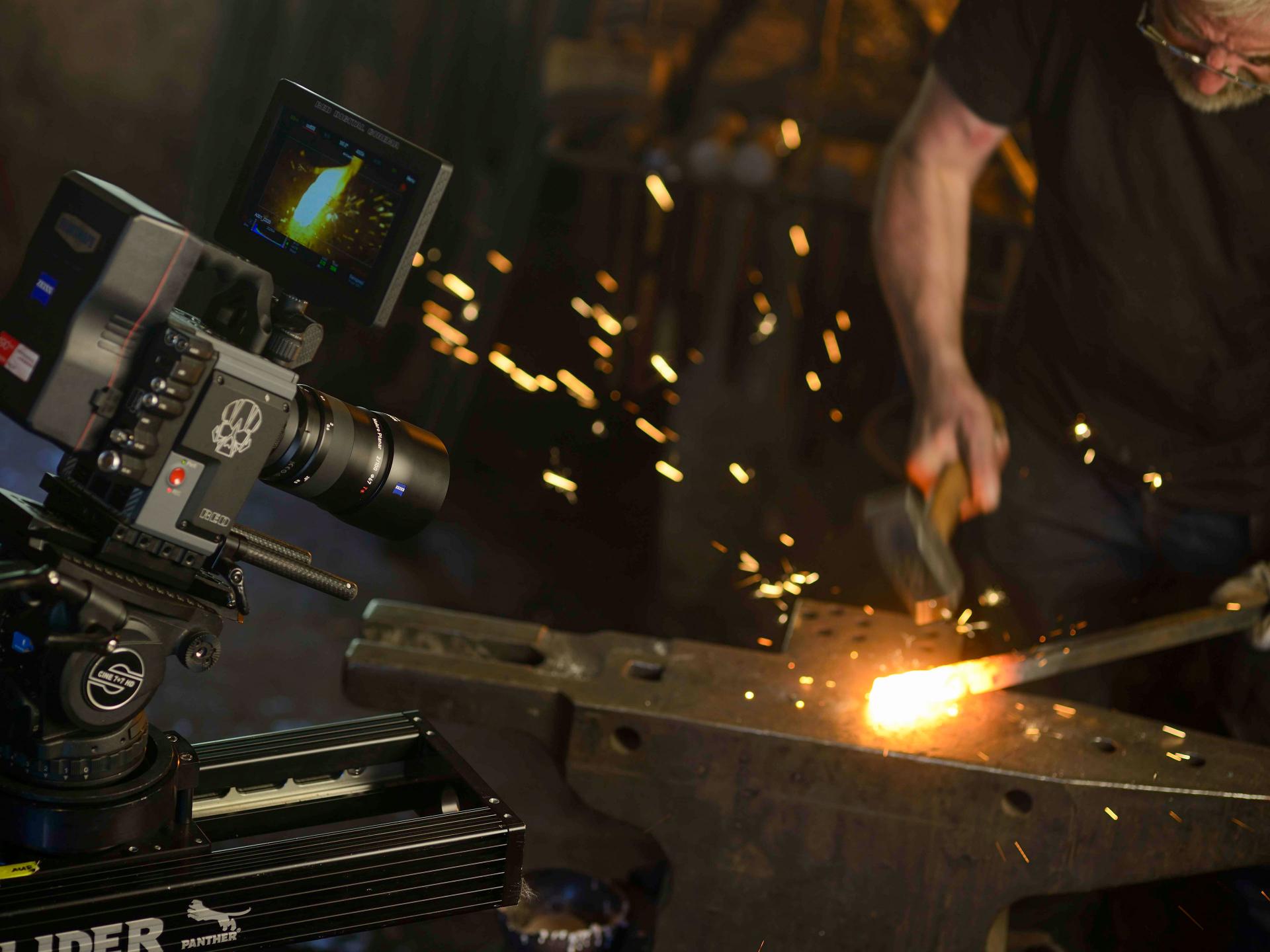 A professional video camera equipped with a ZEISS lens captures footage of a blacksmith hammering glowing hot metal, with sparks flying.