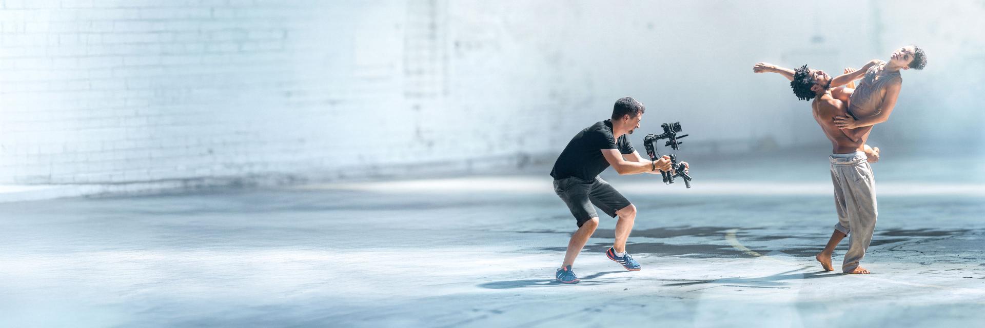A videographer films two contemporary dancers performing in a spacious, well-lit industrial-style setting using a camera rig equipped with ZEISS lenses.