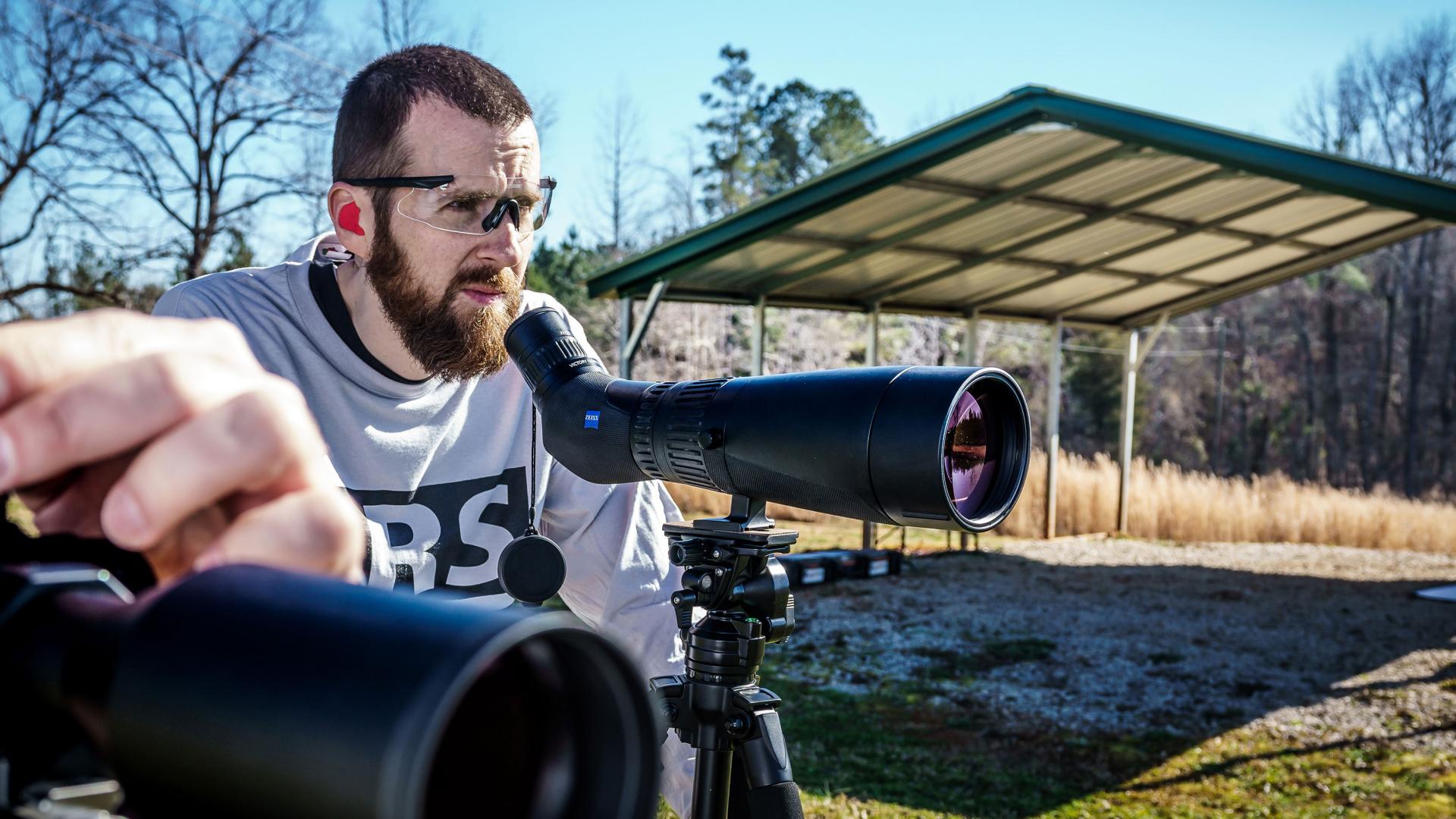 A man outdoors uses a ZEISS spotting scope mounted on a tripod while another ZEISS optical device is positioned in the foreground.