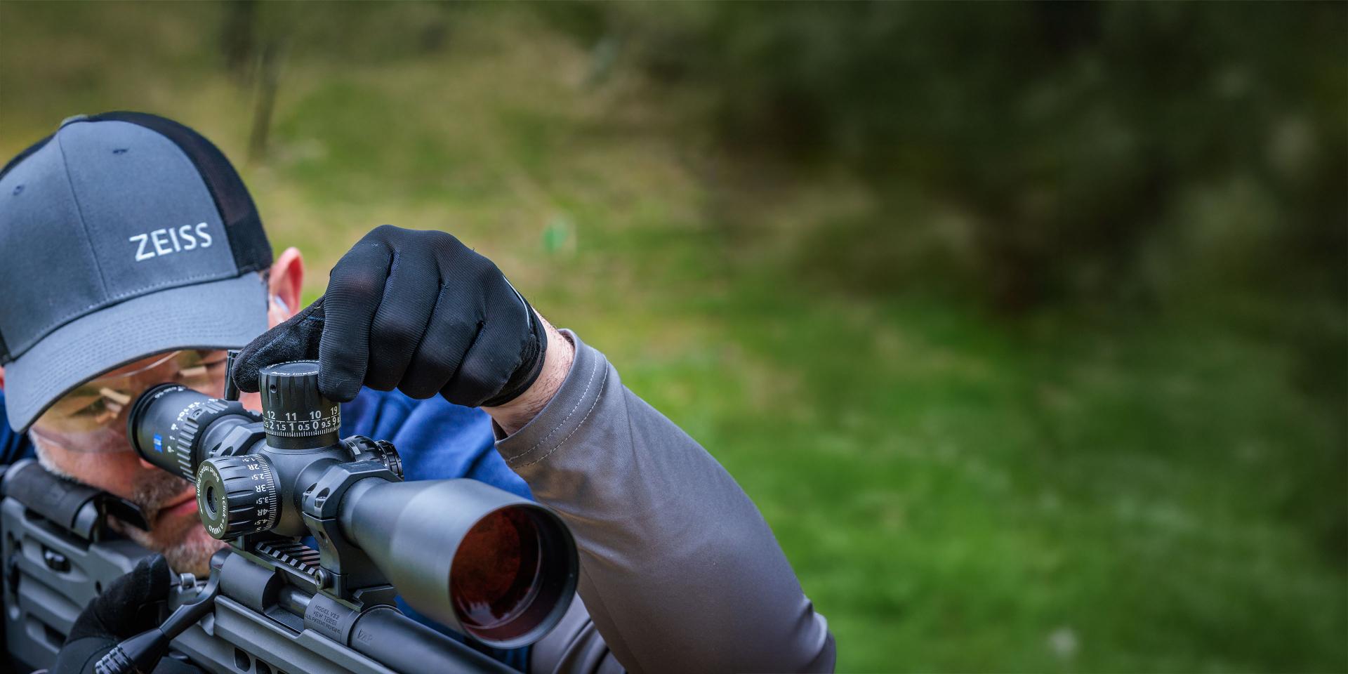 Man adjusting the scope of a high-precision rifle equipped with a ZEISS optic, outdoors in a green, wooded area.