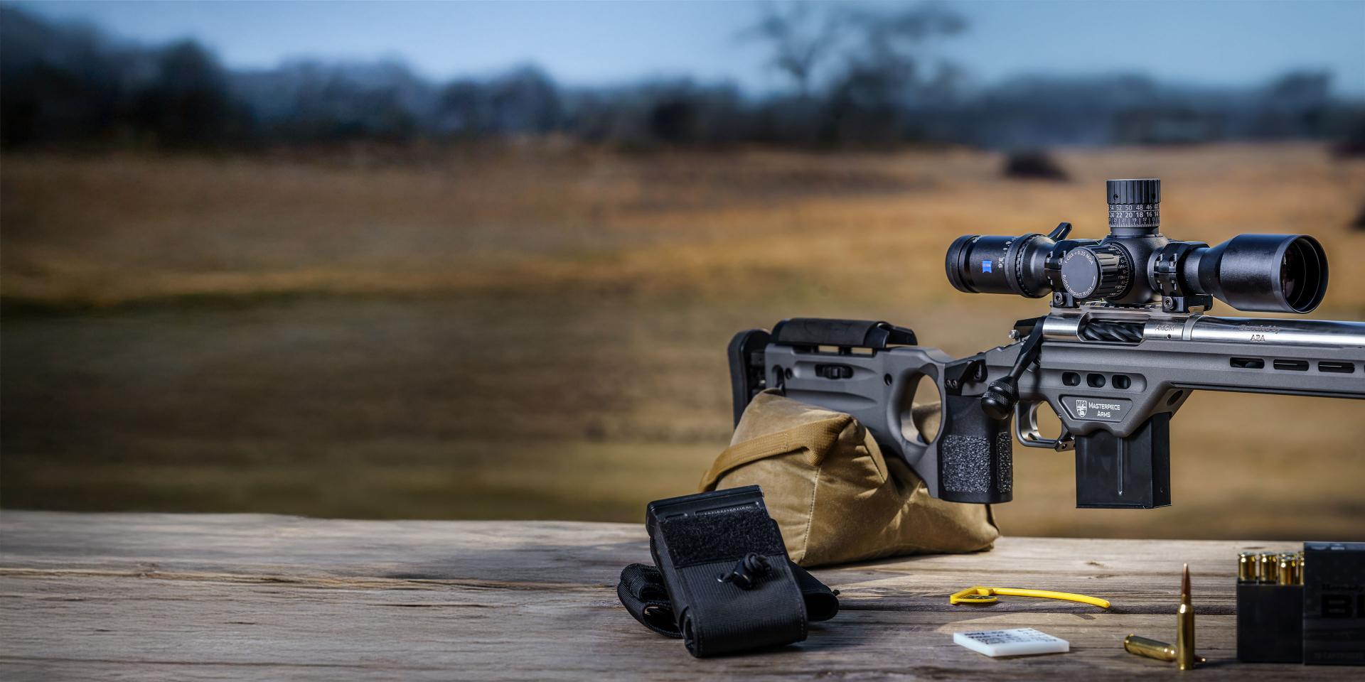 A precision rifle equipped with a ZEISS riflescope rests on a shooting bench with various shooting accessories and ammunition in the foreground, overlooking an open field.