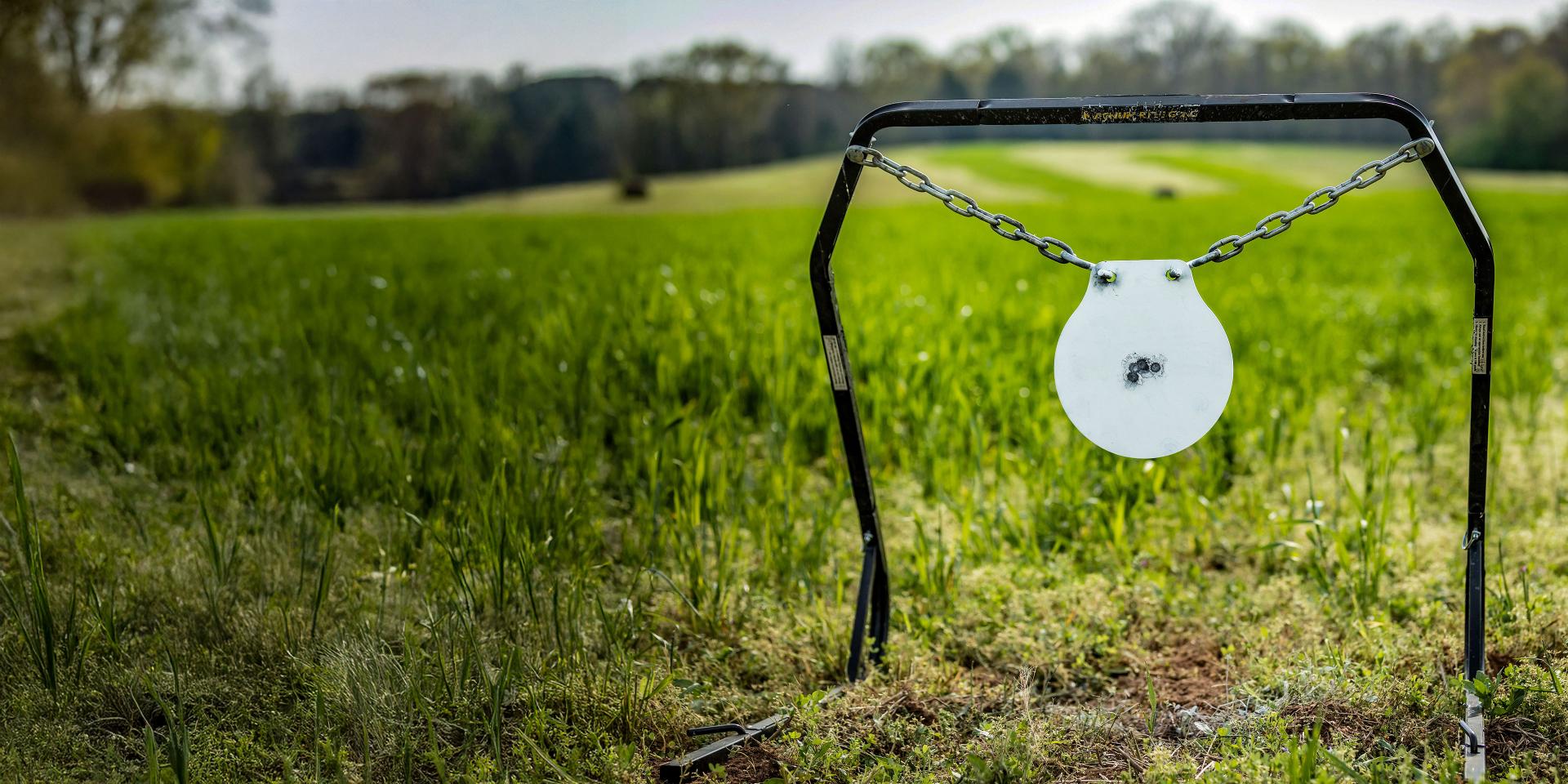 Hanging metal target with multiple bullet impacts suspended by chains between two black stands in a grassy outdoor field.