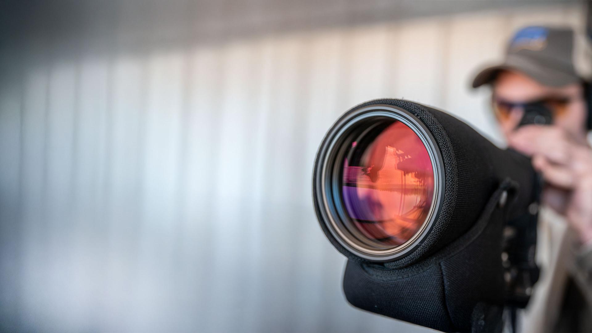 A person using a ZEISS spotting scope with a large lens reflecting light in an outdoor setting.