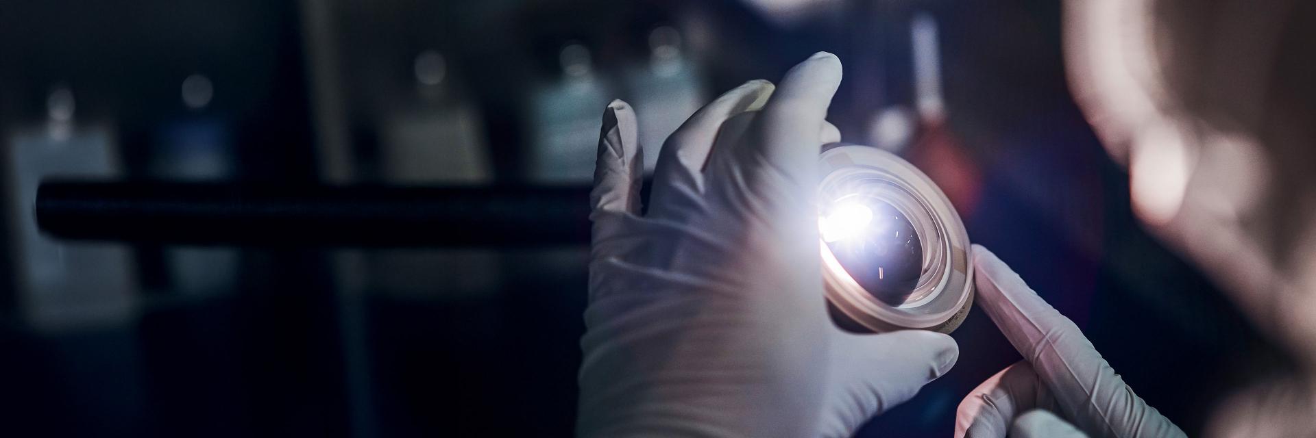 Gloved hands inspecting a ZEISS optical lens under a focused light in a laboratory setting.