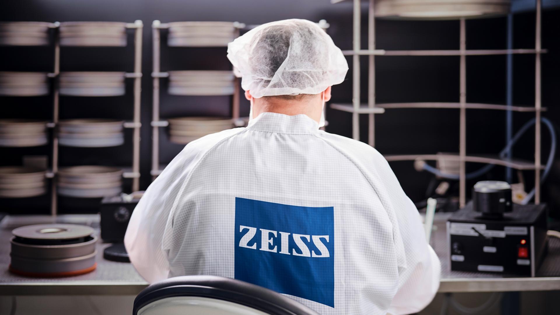 A technician wearing a lab coat and hairnet with the ZEISS logo works in a cleanroom environment surrounded by precision equipment and materials.