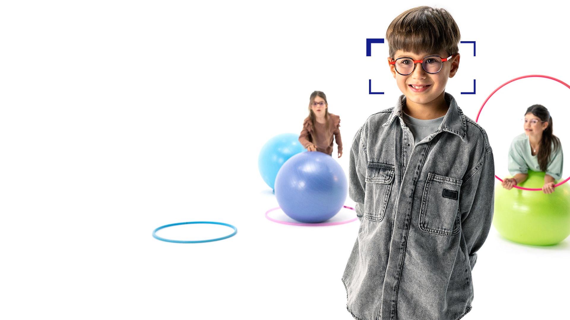 Un garçon aux cheveux marron portant des lunettes rondes et des verres ZEISS MyoCare est debout au premier plan et sourit pour la photo. À l’arrière-plan, deux filles portant des verres ZEISS MyoCare jouent avec des cerceaux et des ballons de gymnastique.
