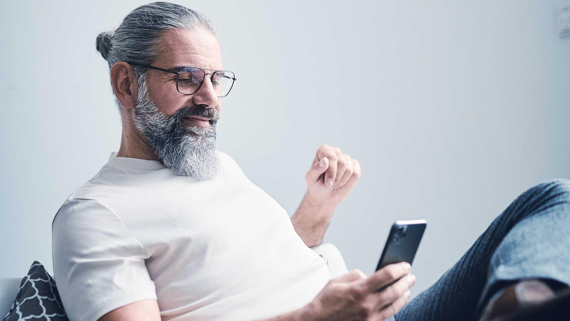 Un homme aux cheveux gris et à la barbe épaisse porte des lunettes, il est assis sur le canapé et regarde son smartphone