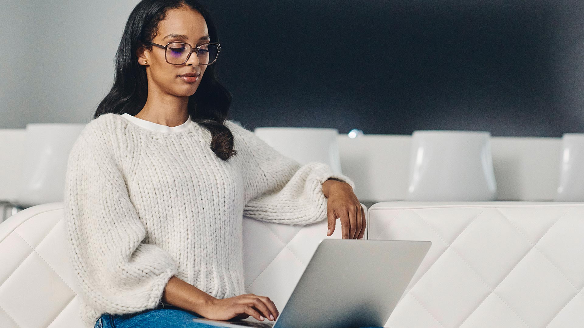 Femme aux cheveux foncés portant des lunettes foncées et un pull blanc regarde un ordinateur portable.