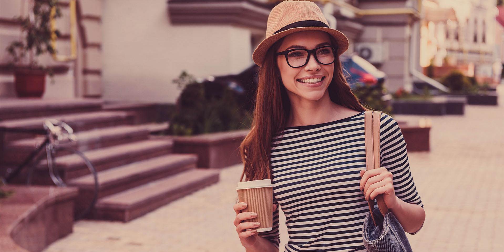 A young woman with a cap and glasses carries a coffee mug (right side) and a bag on her left shoulder