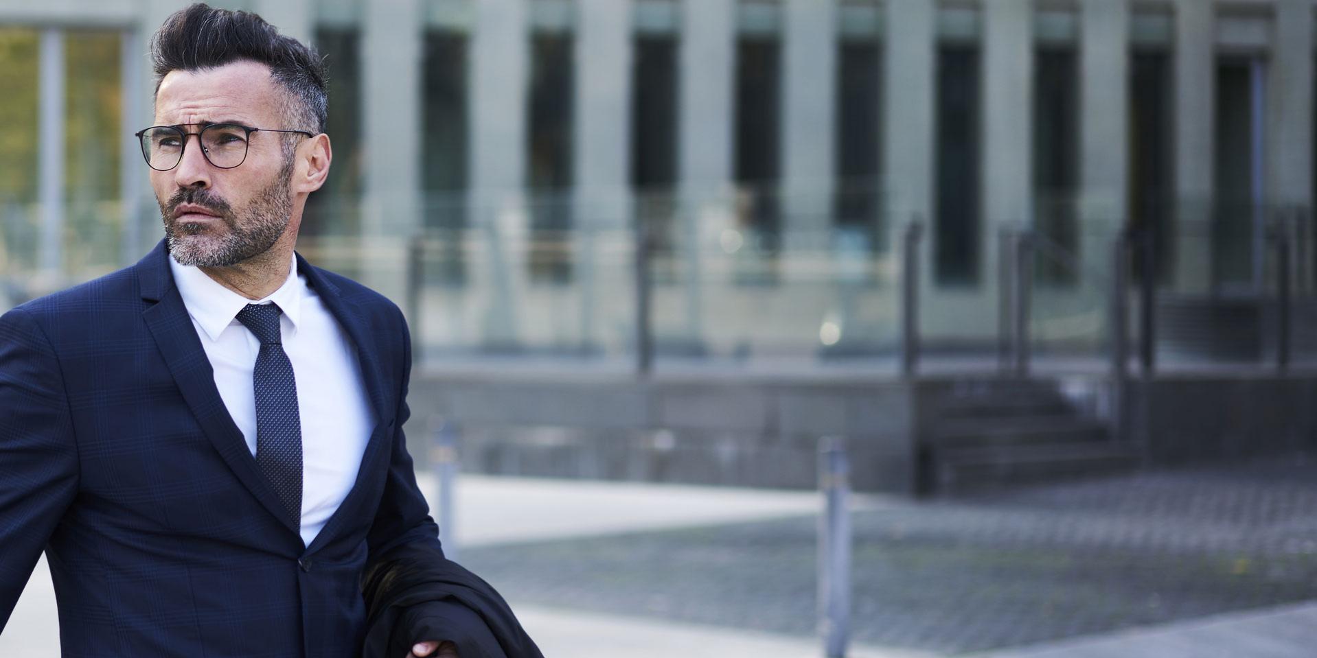 Un avocat barbu et confiant, portant des lunettes à la mode, traverse la rue en passant devant des bâtiments d&apos;architecture moderne abritant des sièges sociaux dans le quartier des affaires d&apos;une ville, en se dépêchant de se rendre à un rendez-vous.