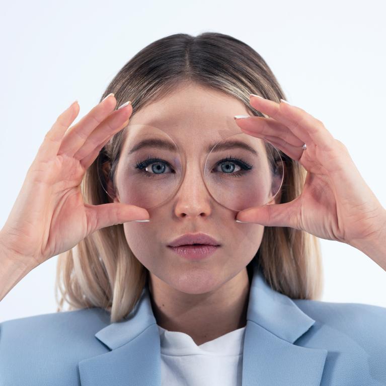 Une jeune femme blonde tient des verres devant ses yeux pour montrer un look esthétique sans effets étranges sur les yeux grâce aux verres unifocaux ZEISS ClearView.
