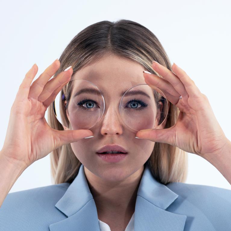 Une jeune femme blonde tient des verres devant ses yeux pour montrer l’effet « fisheye » causé par les verres négatifs épais.
