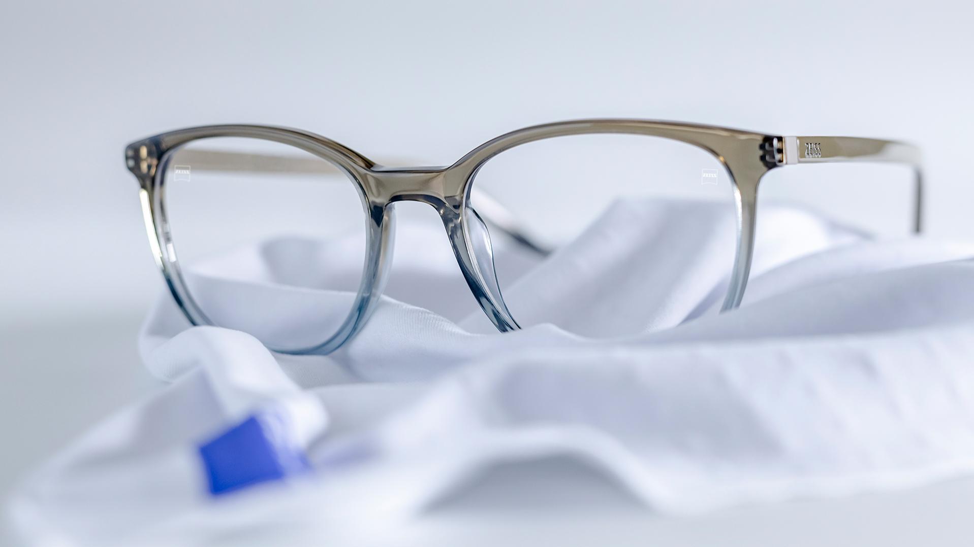 Une paire de lunettes avec une monture gris-bleu et des verres ZEISS avec un traitement DuraVision® est placée sur un chiffon microfibres blanc.