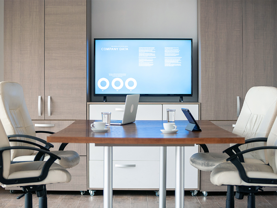 Vue depuis la chaise d’un bureau de cadre supérieur. Un écran est monté sur un mur à quelques mètres. On voit une petite table de réunion entourée de chaises de bureau et sur laquelle sont posés des ordinateurs portables.