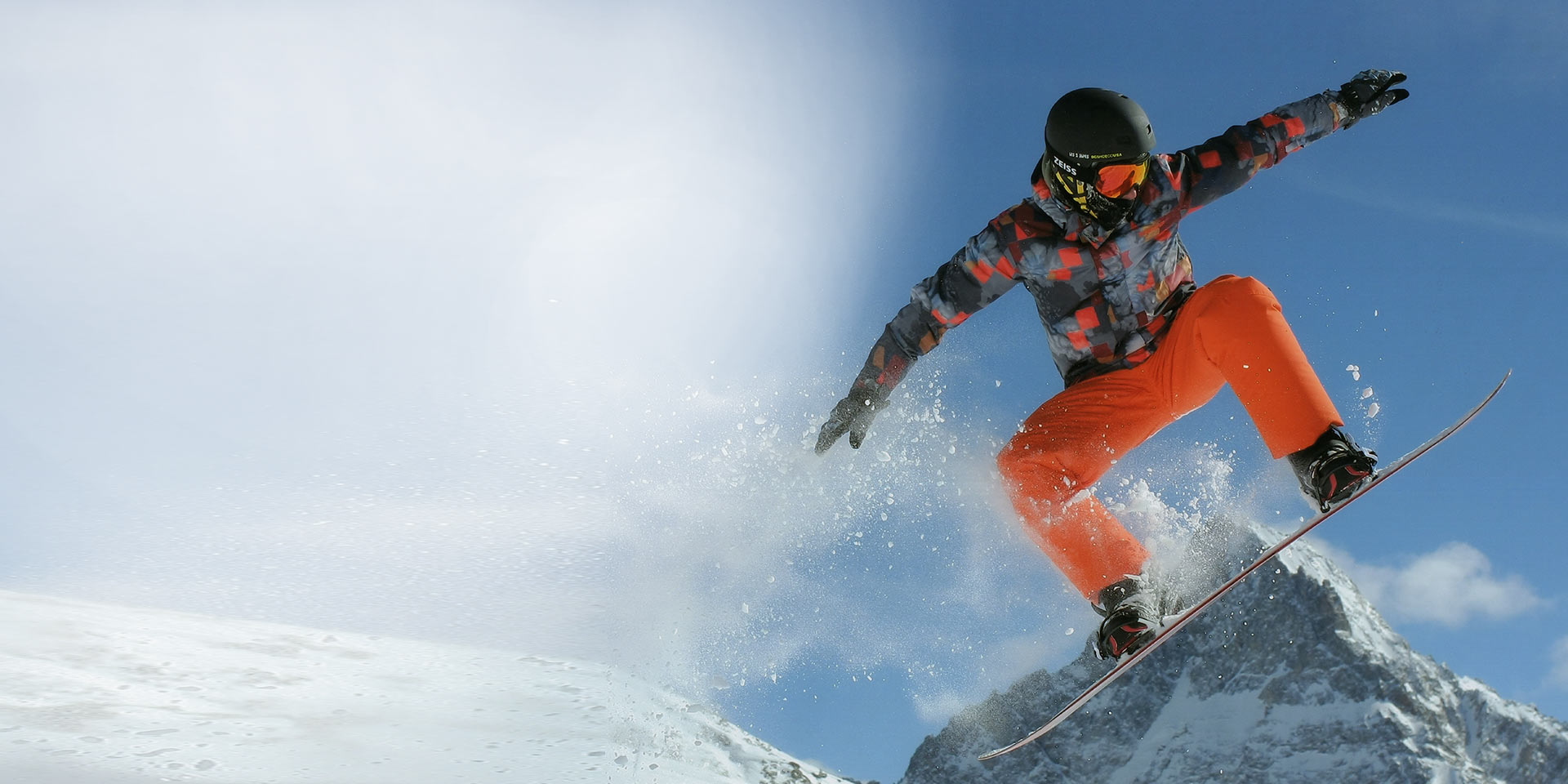 Un homme saute avec un snowboard, porte un casque de ski noir et des lunettes de neige ZEISS avec technologie interchangeable pour la protection.